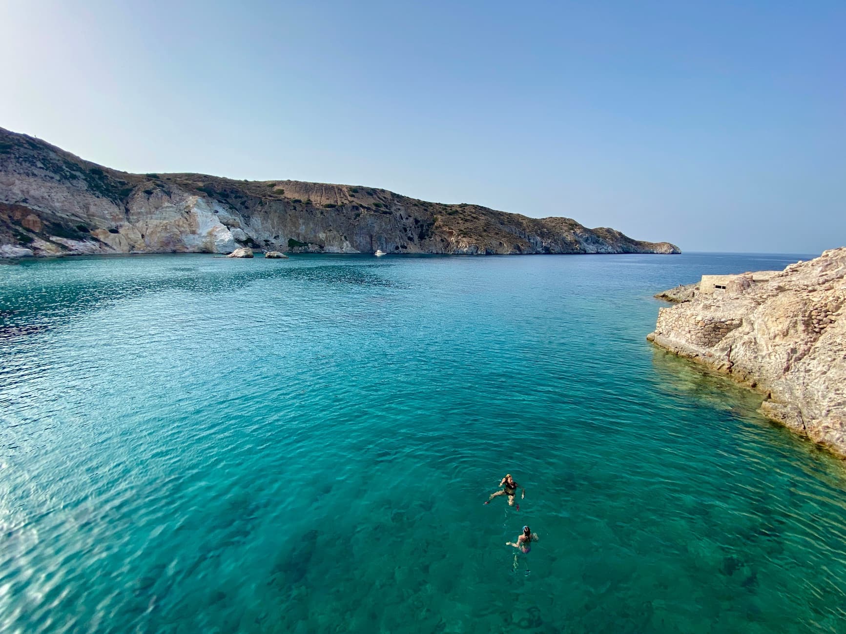 People swimming in clear turquoise water below a rocky coastline on Milos