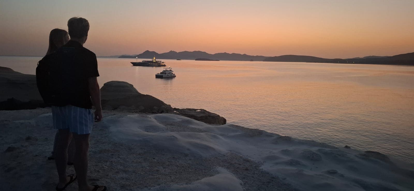 Couple standing on Sarakiniko as cruise ships pass at sunset, Milos